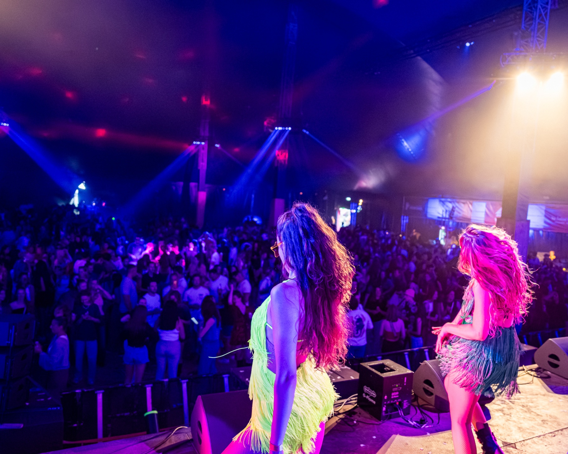 Twee danseressen in neon franjekostuums op het podium boven een vol publiek